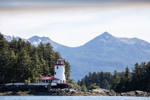 A white lighthouse with a red roof stands by the water, surrounded by trees and mountains in the background.