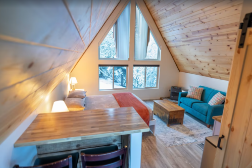 Cozy A-frame cabin interior with a bed, sofa, wooden table, and large windows showcasing a snowy landscape outside.