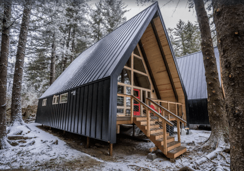 A modern A-frame cabin nestled in snowy woods, featuring a wooden deck and large windows.