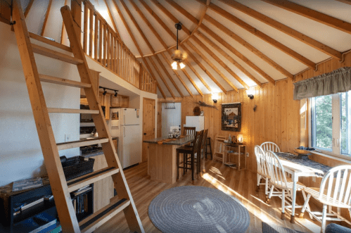 Cozy interior of a wooden cabin with a loft, featuring a dining area, kitchen, and large windows for natural light.
