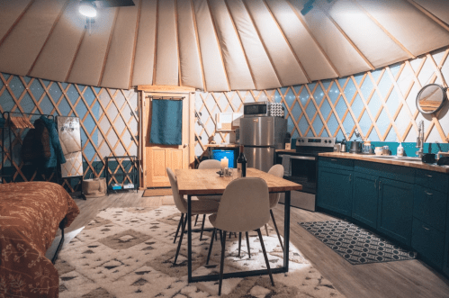 Cozy yurt interior featuring a dining table, kitchen area, and a bed, with warm lighting and rustic decor.