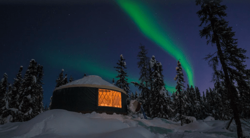 A cozy yurt illuminated at night, surrounded by snow and trees, under a vibrant display of the northern lights.