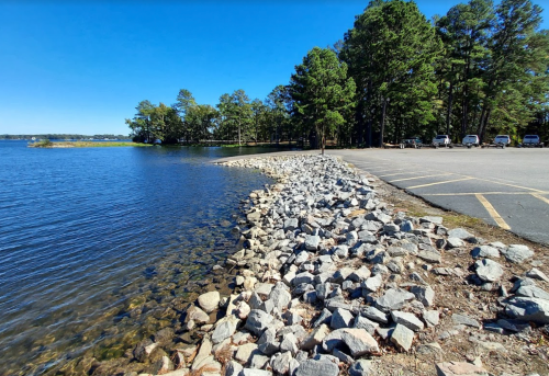 A rocky shoreline beside a calm lake, with trees in the background and a parking area nearby under a clear blue sky.