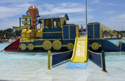 Colorful water play structure resembling a train, featuring slides and water features, set against a blue sky.