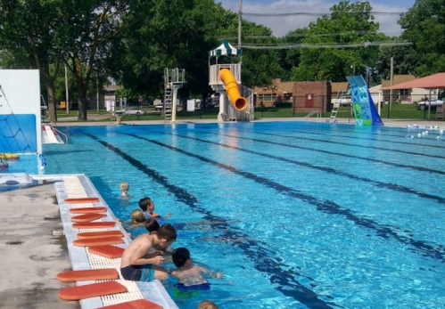 A sunny outdoor swimming pool with a water slide, children playing in the water, and lounge chairs along the edge.