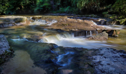 A serene river flows over rocky terrain, creating small waterfalls surrounded by lush greenery.