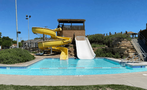 A bright outdoor pool area featuring two yellow water slides and a white slide, surrounded by greenery and blue skies.