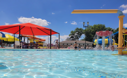 A sunny pool scene with colorful water slides, a splash pad, and shaded seating areas under bright blue skies.