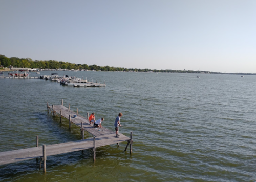 A wooden dock extends into a calm lake, with two children and an adult fishing, surrounded by boats and greenery.
