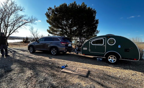 A car parked next to a green teardrop trailer, with a bike attached, near a lake and trees under a clear blue sky.
