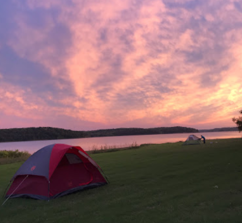 A serene lakeside campsite at sunset, featuring two tents and a colorful sky with pink and purple hues.