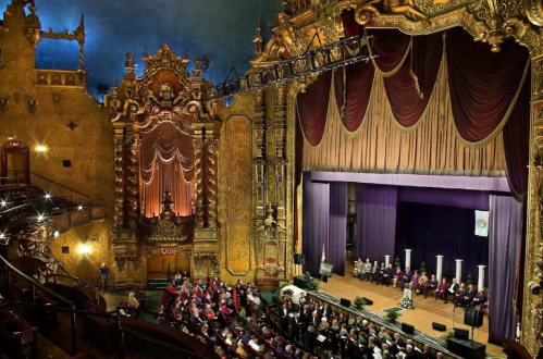 A grand theater interior with ornate decorations, featuring a stage and an audience seated in rows.
