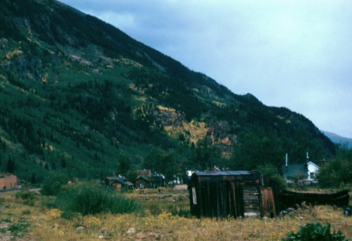 A scenic view of a mountainous landscape with a rustic cabin and greenery under a cloudy sky.