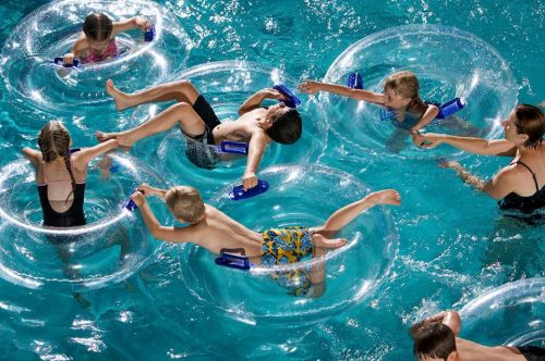 Children and an adult play in a pool, floating on inner tubes and splashing water around.