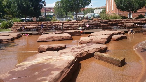 Children play in a shallow water area with large rock formations and a gentle waterfall in a sunny park setting.
