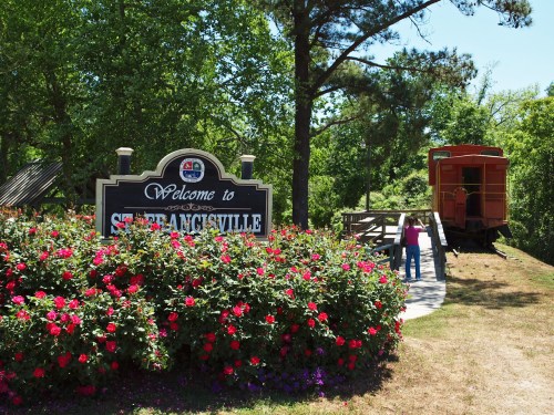 Welcome sign for St. Angsville surrounded by colorful flowers, with a vintage train car in the background.