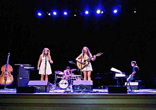 A live music performance featuring two singers, a drummer, and musicians on guitar and piano on a dimly lit stage.