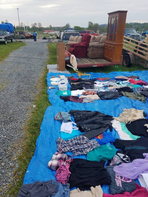 A blue tarp on the ground displays various clothes for sale, with a couch and vehicles in the background.