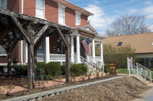 A brick building with a porch, American flag, and landscaped front yard, set against a clear blue sky.