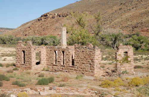 Ruins of a stone building surrounded by sparse vegetation and mountains under a clear blue sky.