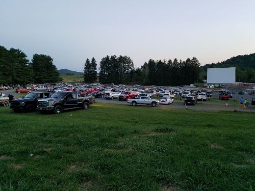 A drive-in movie scene with numerous parked cars and a large screen in the background, surrounded by trees.