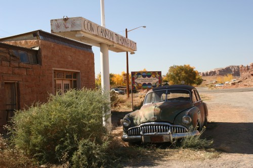 An old, rusty car sits beside a weathered sign and a stone building in a deserted area with sparse vegetation.