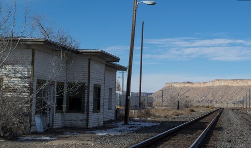 Abandoned building beside railway tracks, with a rocky cliff in the background under a clear blue sky.