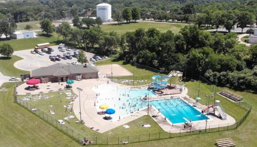 Aerial view of a community pool with slides, surrounded by green grass and trees, and a parking lot nearby.