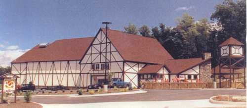 A large, rustic building with a steep roof, featuring wooden beams and a stone tower, set in a wooded area.