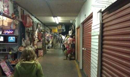 A narrow market corridor lined with various stalls and merchandise, with a person walking in the foreground.