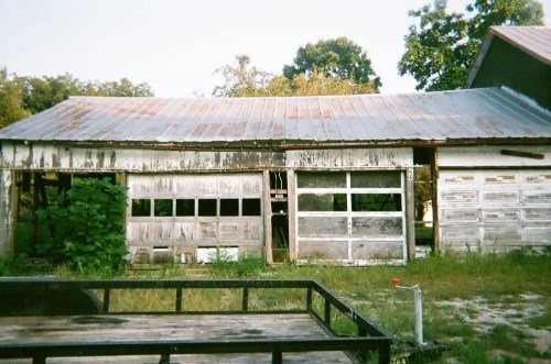 An old, weathered garage with peeling paint and overgrown vegetation, set against a backdrop of trees.