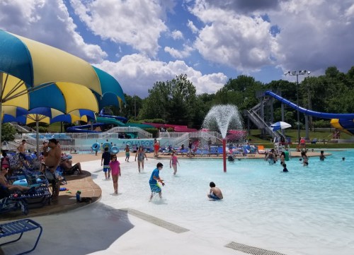 A lively water park scene with children playing in a pool, water slides, and colorful umbrellas under a cloudy sky.