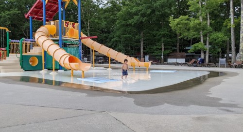 A child stands in shallow water near colorful water slides at a park, surrounded by trees and lounge chairs.