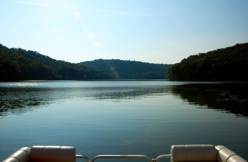 A serene lake view surrounded by hills, with calm water reflecting the clear sky. A boat's edge is visible in the foreground.