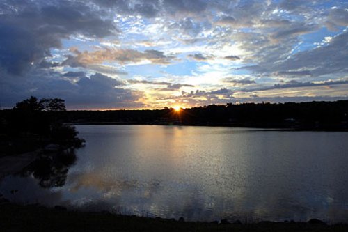 Sunset over a calm lake, with colorful clouds reflecting on the water's surface and trees lining the shore.