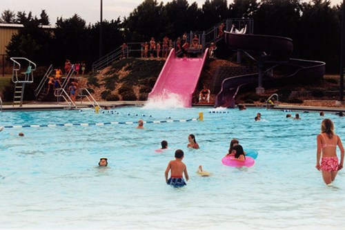 A busy swimming pool with children playing, a pink water slide, and people enjoying the water on a sunny day.