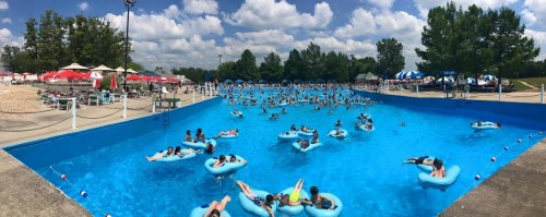 A crowded outdoor pool with many people floating on inner tubes under a bright blue sky and scattered clouds.