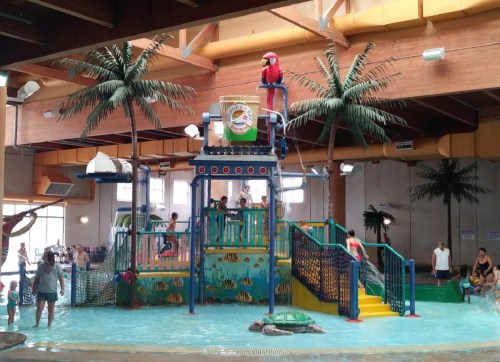 Indoor water park with a colorful play structure, palm trees, and children enjoying the water.