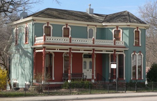 Victorian-style house with a blue exterior, red accents, and a wraparound porch, surrounded by a fence and trees.
