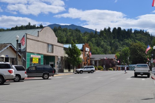A quiet small town street lined with shops, trees, and mountains in the background under a blue sky.