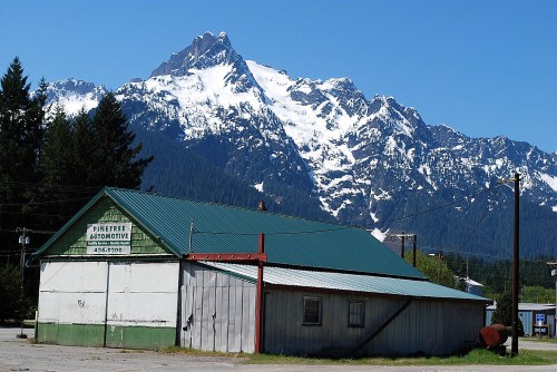 A rustic building with a green roof in front of snow-capped mountains under a clear blue sky.