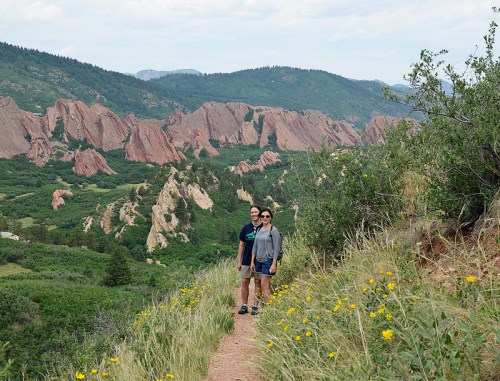 Two hikers stand on a trail surrounded by lush greenery and striking red rock formations in a mountainous landscape.