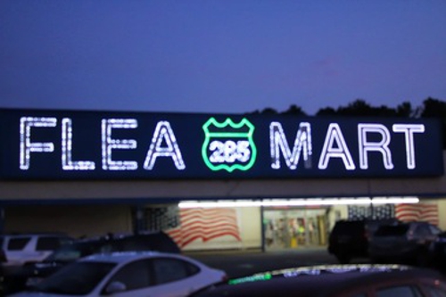 Sign for "Flea Mart" illuminated at night, featuring a highway shield and American flag backdrop.