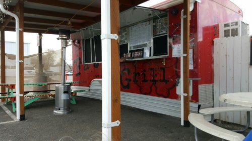 A red food truck with a menu displayed, under a wooden canopy, surrounded by picnic tables in a parking lot.