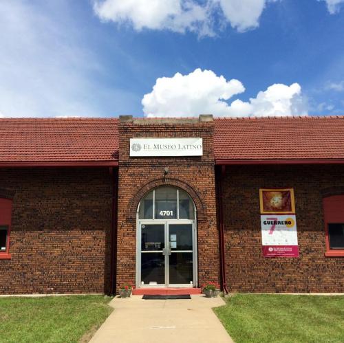 Front view of El Museo Latino, a brick building with a red roof and a sign above the entrance. Blue sky with clouds.