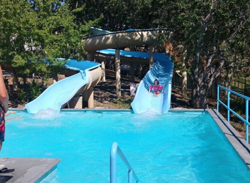 Two water slides empty into a bright blue pool, with a child splashing down one slide on a sunny day.
