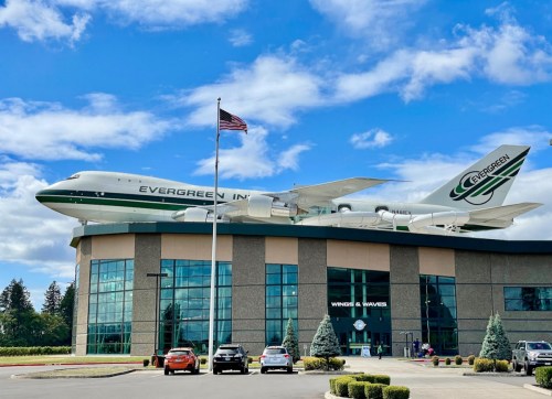 A large airplane model sits atop a building with a flag in front, under a blue sky with scattered clouds.