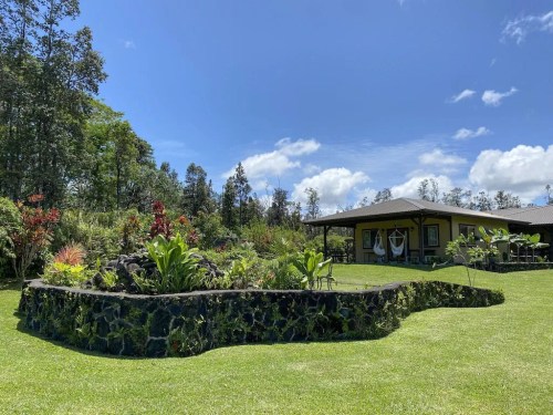 A lush garden with tropical plants surrounds a charming house under a bright blue sky and fluffy white clouds.