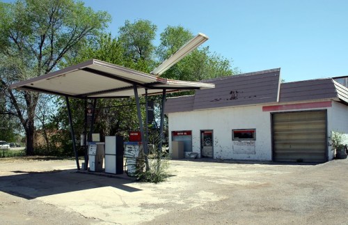 Abandoned gas station with a damaged canopy and a weathered building, surrounded by sparse vegetation.
