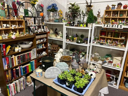 A cozy shop display filled with garden decor, figurines, books, and potted plants, creating a whimsical atmosphere.